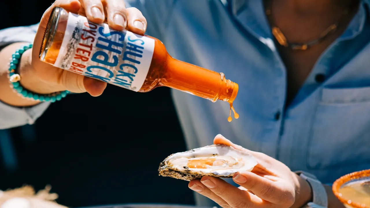 Photo of person holding oyster and Shuckin' Shack hot sauce bottle