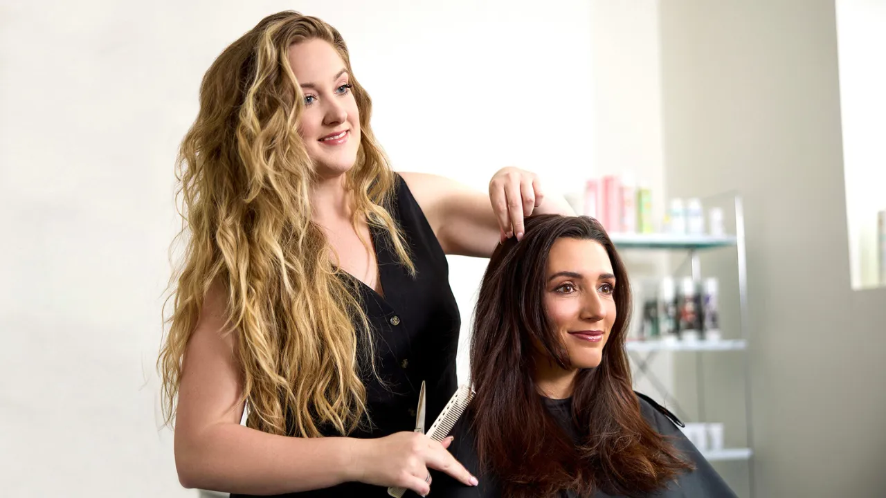 Blonde beauty professional holds comb and shears while styling brunette client's hair