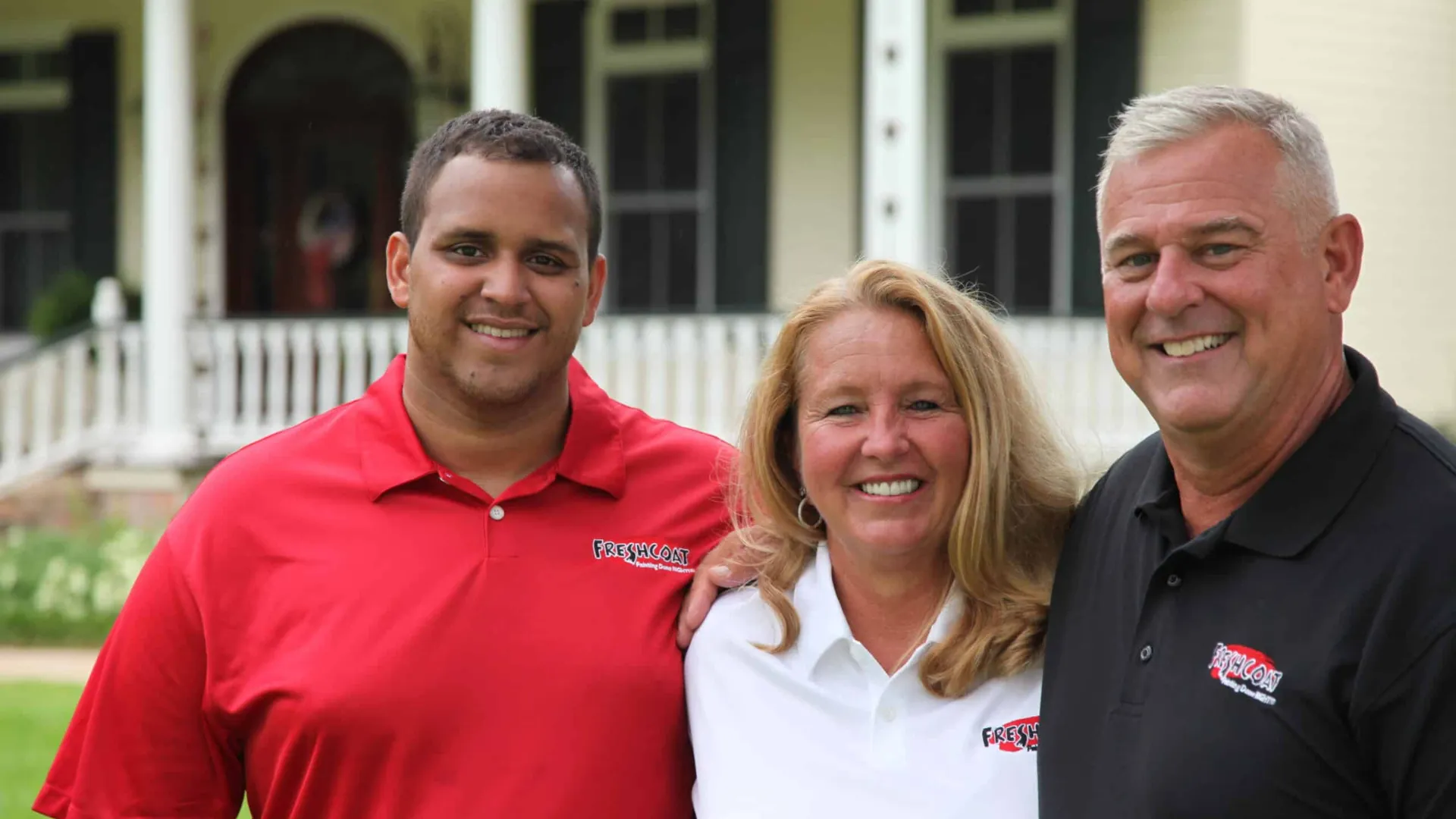 Three Fresh Coat Painters representatives stand, smiling, outside a home in Fresh Coat polo shirts