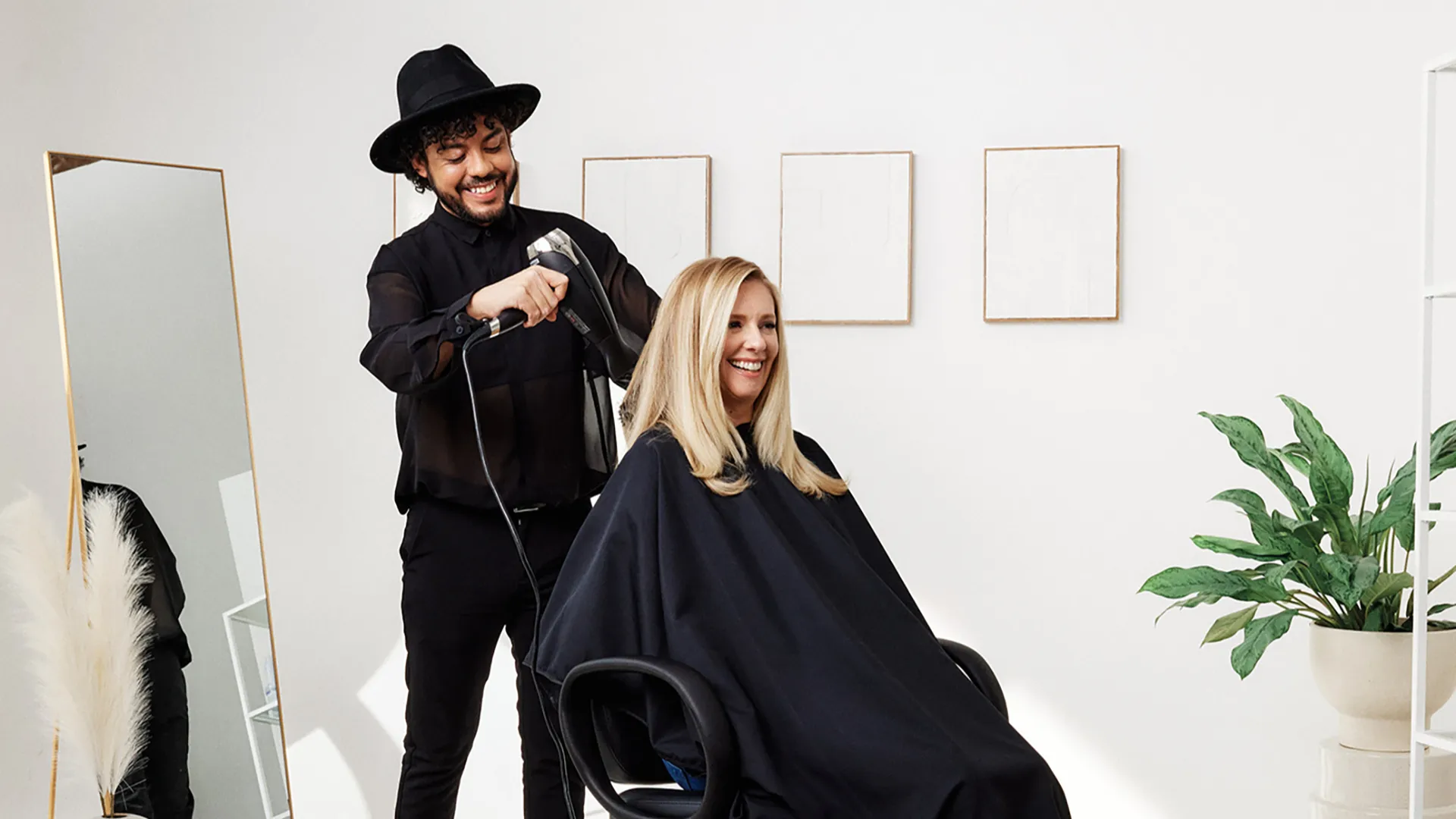 photo of a hairstylist, wearing all black, blow drying a client's long, blonde hair