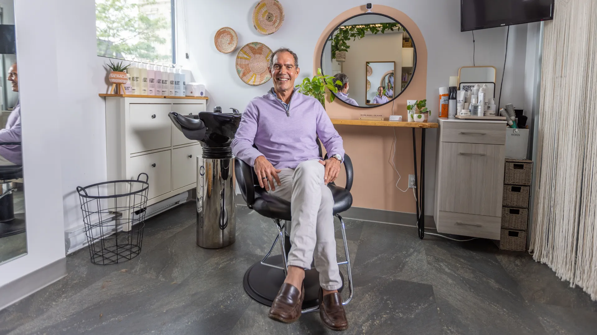 person, wearing purple quarter zip sweater and khakis, sits in salon chair, smiling at camera